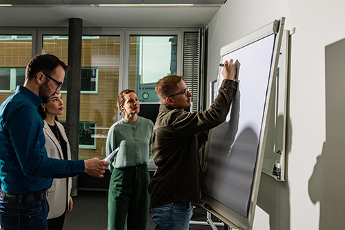 Ein Mann schreibt etwas auf ein Flipchart. Um ihn herum stehen mehrere Personen