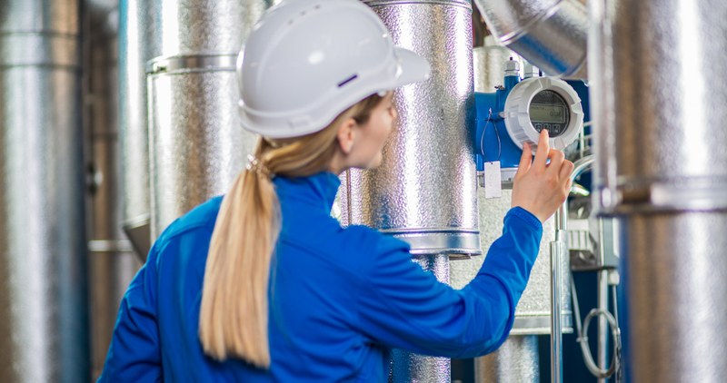 Eine Mitarbeiterin von Endress+Hauser Flow liest am blauen Durchflussmessgerät in der Mitte einer Rohrleitung Messdaten ab.