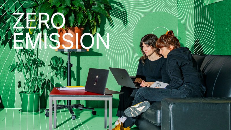 Zwei Frauen sitzen nebeneinander und schauen in einen Laptop. Der Hintergrund des Bildes ist grün. Auf dem Bild steht der Zero Emission