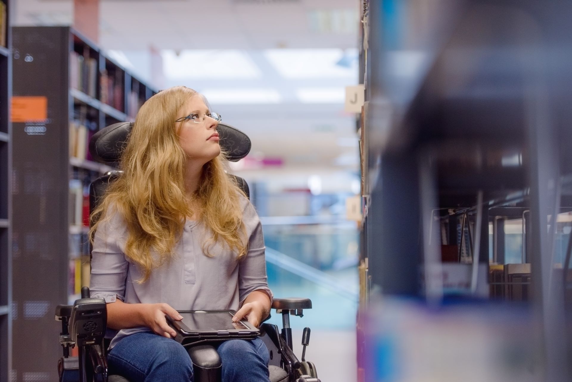 Eine Frau in einem elektronischen Rollstuhl hält ein Tablet in der Hand und blickt in ein Bibliotheksregal