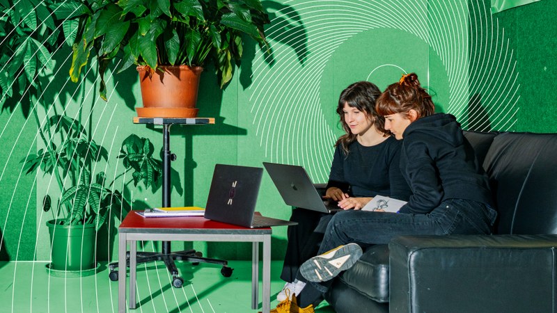 Zwei Frauen schauen auf einen Laptop gemeinsam. Der Hintergrund des Bildes ist grün.