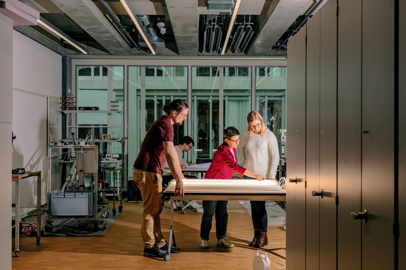 Ein Mann und zwei Frauen stehen an einem Tisch. Auf dem Tisch liegt ein hell leuchtendes Flachbettgerät. Eine Frau zeigt etwas auf dem Gerät.