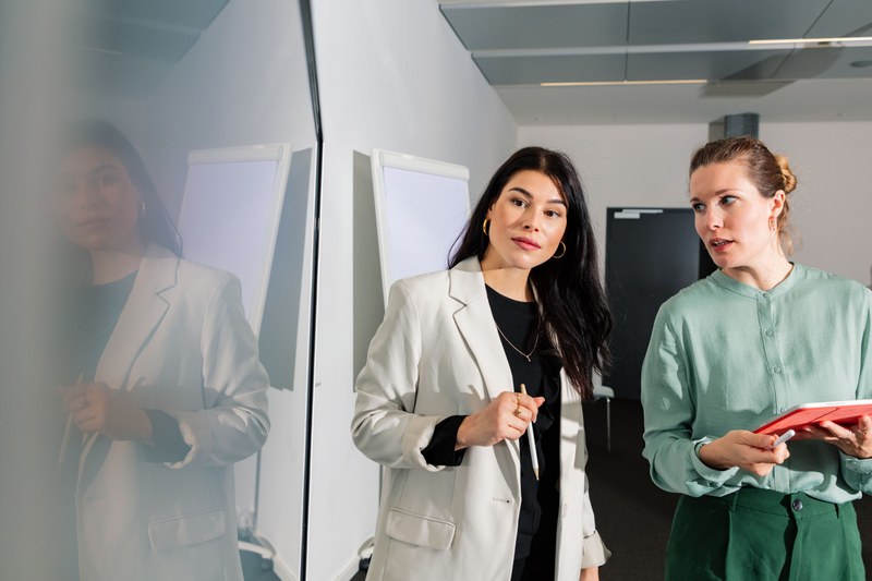 Zwei junge Frauen in Business-Outfit. Eine Frau hat einen Stift für ein Whiteboard in der Hand, die andere in Tablet