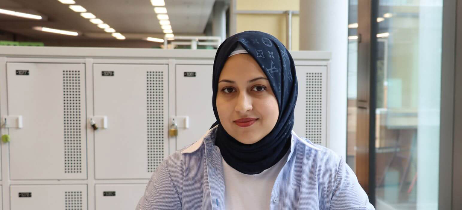 Elham Sherifi sitzt im Campus an einem Tisch und lernt. Sie hat ein dunkelblaues Kopftuch an und sieht in die Kamera