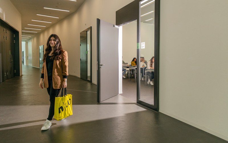 Eine junge Frau tritt lächelnd aus einem Unterrichtsraum der FHNW. Sie hat eine gelbe Tasche mit FHNW-Logo in der Hand.