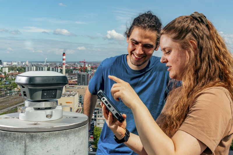 Ein Mann und eine Frau auf einem Dach über dem Basler Industriegebiet. Die Frau hat ein iPad mit dem sie die Daten eines Geomatik-Sensors auswertet, der auf dem Dach installiert ist