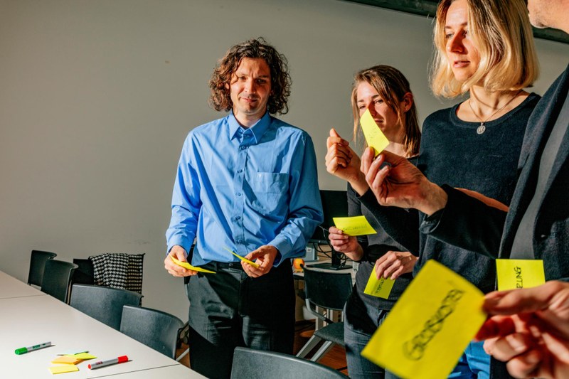 Teilnehmende einer Weiterbildungsveranstaltung stehen um einen Tisch und halten Post-It-Zettel in den Händen